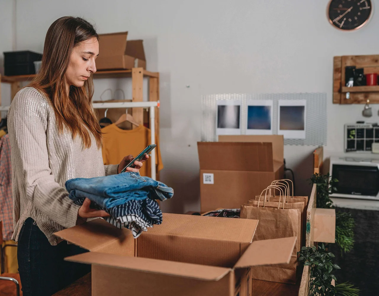 Women looking at phone hold a pile of fold clothes ready to put in a box
